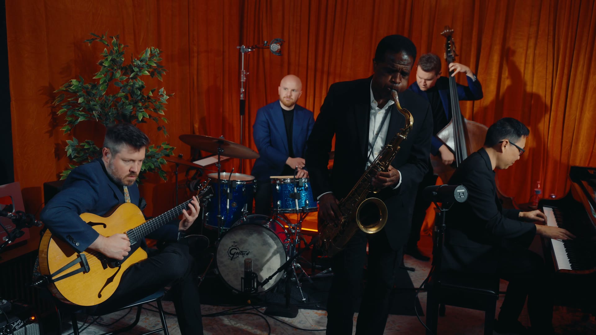The full Sessions With Shaw band live in the studio: Rickey Woodard on tenor saxophone with guitar, drums, bass, and piano against the orange curtain